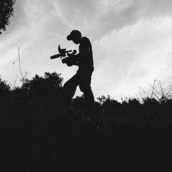 Production still from Silent Beauty. Low-angle shot of a cameraperson walking with equipment in hands, surrounded by nature. Black and white overshadowed shot.