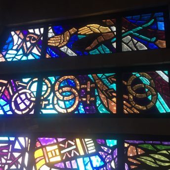 Still from Mama Bears. A stained glass window of purples, greens, and golds. There are multiple linked gender symbols, hands, and letters that read, "ran" and "za".