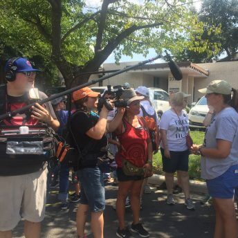 Production still from Mama Bears. There is a crowd of people, in the foreground are two film crew members. One crew member has a camera and the other has a boom microphone, speaking to and filming a woman.