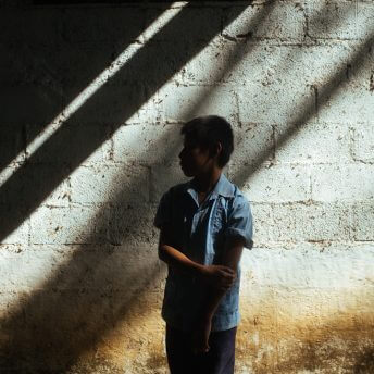 A young boy with a blue shirt looks away from the camera. He stands in front of a bricked wall. Lines of light reflect on the wall and the boy.