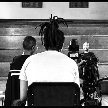 Still from Milisuthando (working title). Two crew members are sitting infront of and filming Milisuthando Bongela, who is seated in front of a wooden stadium-style bench seating. The photo is in black and white.