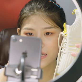 Still from Ascension. A woman is making a video with her cellphone, she is holding a tennis shoe close to her face while speaking.