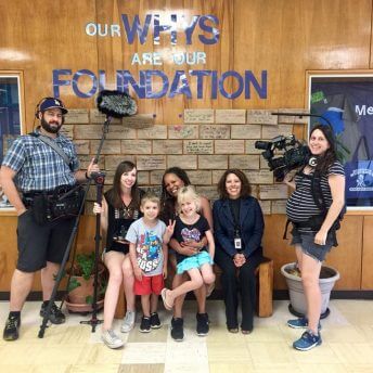 Production still from Mama Bears. Seven people are in front of a wall that says, "Our Whys Are Our Foundation". There are three crew members with camera equipment. Two crew members are standing and one is sitting on a bench with two women and two children.