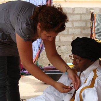 Farah Kassem bent over affixing a microphone onto a person's shirt. Color photograph.