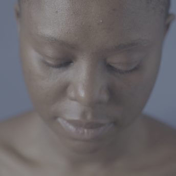 Still from Milisuthando (working title). A close-up, downward angle of a woman who is looking down and away from the camera. The background is blue.