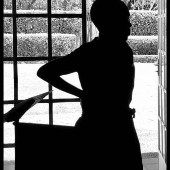 Still from Milisuthando (working title). A person is standing with their hands on their hips in front of a windowed door open to an outside garden area. Backlit, black and white photo.