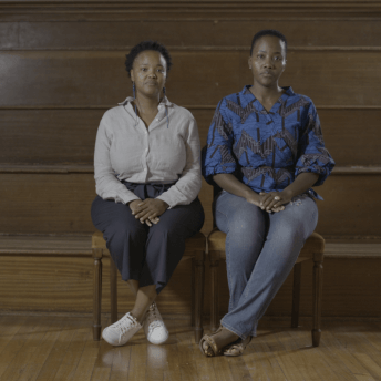 Still from Milisuthando (working title). Two women are sitting in front of a wooden stadium-style bench seating with their hands in their laps and ankles crossed.