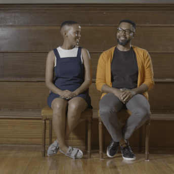 Still from Milisuthando (working title). A man and a woman sit in two wooden chairs in front of a wooden stadium-style bench seating. They are both smiling as the woman looks at the man.