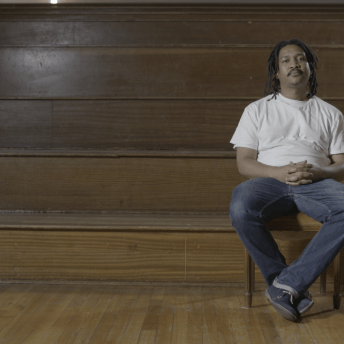 Still from Milisuthando (working title). A man sits in front of a wooden stadium-style bench seating with his hands in his lap and ankles crossed.