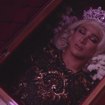 Still from #Mickey. The top half of a wooden casket is open, revealing a woman wearing a black and gold sequin dress and crown. There are white and red flowers by her head.