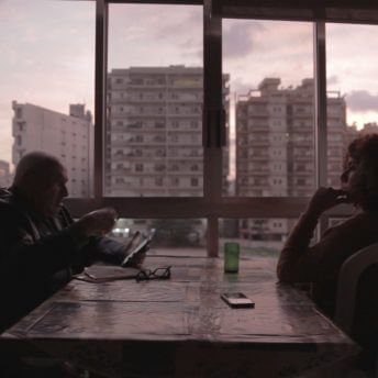 Farah Kassem and her father sit across from each other at a table. Farah's father Mustapha is looking at his phone. In the background, tall buildings can be seen through the large window. Color photograph.