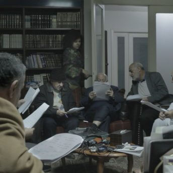 A group of men sits on couches holding papers in front of them. Farah Kassem is walking in the background next to a bookshelf. Color photograph.
