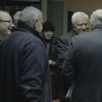 A group of men in suits crowds around two men shaking hands. Color photograph.