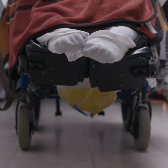 A photo of a person sitting in a wheelchair, only their feet are visible and they are wearing white socks. A red blanket over their lap.
