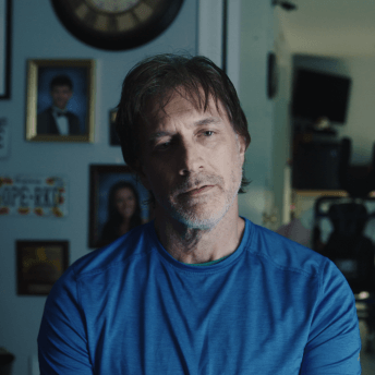 Still from Storming. A man in a blue t-shirt is sitting in front of the camera, he looks away. In the background there are portraits hanging on the walls.