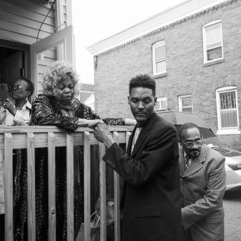 Black and white photo. A man in the foreground is holding the hand of an older woman who is standing on a wood porch. The woman has curly grey hair and is wearing a breathing tube on her nose. There are other men and women surrounding them. In the background is a brick building behind them.