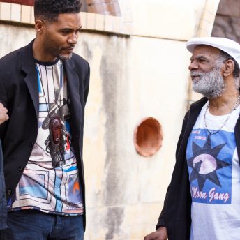 Three people converse outside of a building. There are two men–one middle aged and one elderly with a white beard –and a woman.
