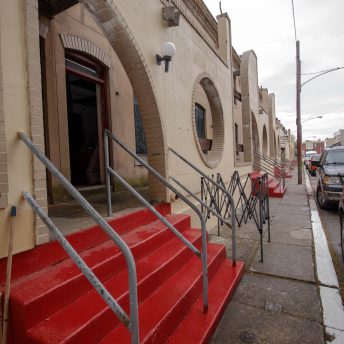 Street view of a beige-exterior building. The building has a set of stairs painted bright red, and a façade wall with a round hole.