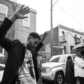 Black and white photo. A man in the foreground is lifting his arms up, and his eyes are closed. To his right are a group of women and men in formal wear. Two women shelter under an umbrella.