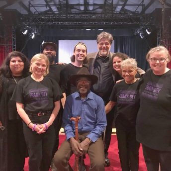 Production still from Frank Bey: You're Going to Miss Me. Frank Bey–a man in a lilac button-up shirt, brown wide-brimmed hat, and holding a wooden cane–is sitting down, surround by group of eight people , some of whom are wearing a black t-shirt that reads: "Frank Bey: Back in Business." Behind them are partially seen stage curtains and lights.