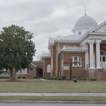 Still from Frank Bey: You're Going to Miss Me. Frank Bey is wearing a dark jacket, wide brimmed hat, and holding a wooden cane. He is standing on a sidewalk in front of a large brick two-story building with elaborate white columns, a tall, triangular entryway covering, and copula. There is a large wooden cross in front of the building, and the building is surrounded by a grassy lawn.