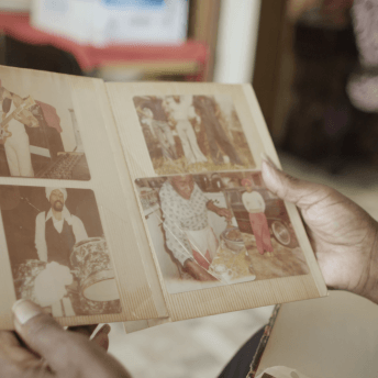 Still from Frank Bey: You're Going to Miss Me. A person is holding open a booklet that displays five photos whose color is faded. Two of the photos portray men playing guitar and drums. A third photo portrays three men in bell-bottom jeans in front of a car. A fourth photo portrays an older woman in an apron preparing food. A fifth photo shows a woman in red bottoms, white shirt, and red headwearp in front of a black car.