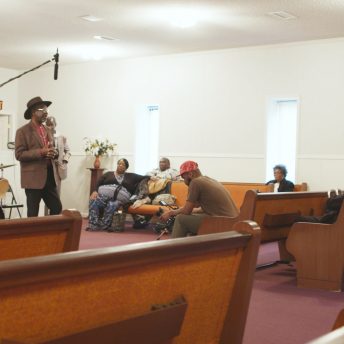 Production still from Frank Bey: You're Going to Miss Me. A man in a brown jacket and wide brimmed hat is addressing a group of people sitting in a wooden pews inside a building. Another is holding a boom mic above him.
