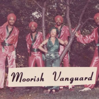 Still from Frank Bey: You're Going to Miss Me. A color photo that appears faded. Six men stand in a semi-circle around a woman dressed in an emerald-green, shiny paintsuit and headwrap. The men are wearing red or green headwraps and red, loose-fit satin shirts and pants. The shirts are trimmed in green satin fabric, and tied with a green satin belt. Black text on a light label reads: "Moorish Vanguard."