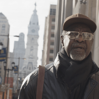 Still from Frank Bey: You're Going to Miss Me. Frank Bey, a man with a salt-and-pepper beard, is wearing gold-frame glasses, brown baseball cap, and black scarf and leather jacket. Behind him tall buildings, street lights, and stoplights are seen slightly out of focus.