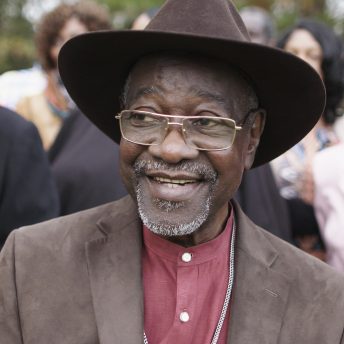 Still from Frank Bey: You're Going to Miss Me. Frank Bey, a man with a salt-and-pepper beard, is wearing gold-frame glasses, a wide brimed brown hat, and a brown suede jacket over a red button-up shirt. Behind him, seen slightly out of focus are other men and women also in formal dress.