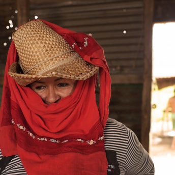 Still from Once Upon a Time in Venezuela. A woman stands near a door to the outside. She is wearing a straw hat with red fabric wrapped around her head, mouth, and neck.