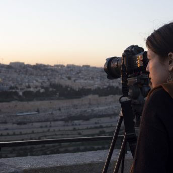 Rachel Elizabeth Seed is in front of a photograph of a city, looking away from the camera, wearing a sequined blouse, and has her hair tightened in a low ponytail. Portrait in black and white.