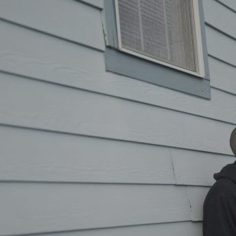 A man leaning against a house, he looks away from the camera.