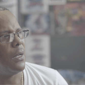 A man wearing a white shirt with eye glasses looks away from the camera to speak with someone out of frame.