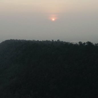 Still from Eskape. A red-orange sun shines over a forest-covered hill, it's rays diffusing out due to the foggy sky.
