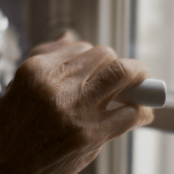 Still from Eskape. Closeup of a woman's hand, holding a window handle as if about to open it. Her hand is in focus, and wrinkles are visible, and her fingernails are painted red. Out of focus, her torso is seen. She is wearing a black sweater and a large silver necklace.