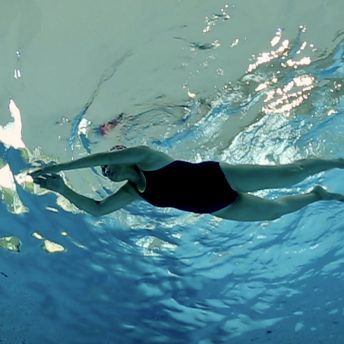 Still from Eskape. Underwater shot of a woman in a one-piece swimsuit and swim cap, swimming with arms crossing in front of her. The water is bright blue and in areas appears silver from the reflection of light on the surface.