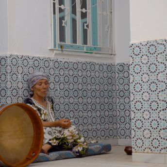 Still from Machtat. A woman in a headwrap sits on a mat on the ground against a wall smoking.