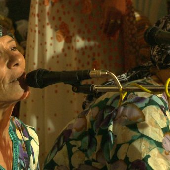 Still from Machtat. Two women in colorful dresses and headwraps. One is singing with a microphone.