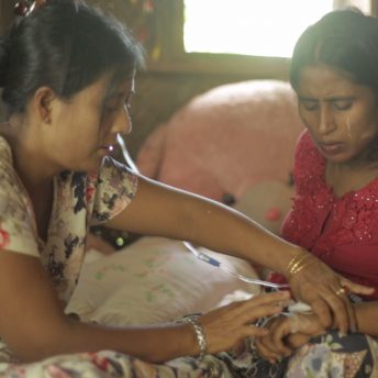 Still from Midwives. Two women are sitting together inside. The focus of both women is on the one touching the wrist and hand of the other.