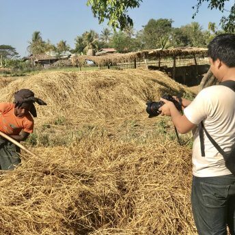 Production still from Midwives. A camera man wearing a backpack is taking video of a young boy moving hay.
