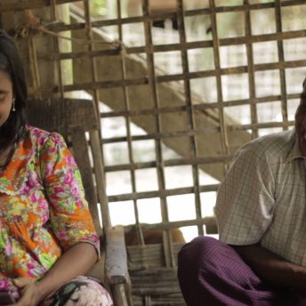 Still from Midwives. A smiling man and woman sit beside each other outside.