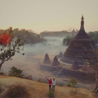 Two people standing atop a hill, the person on the left is waving, with scenery behind them.