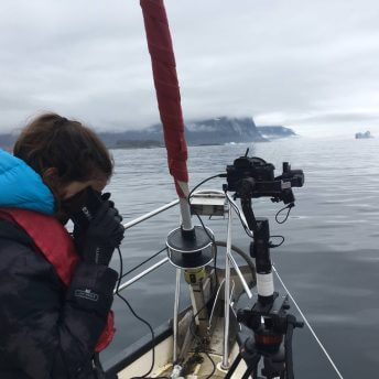 A woman looking down through binoculars standing starboard in a boat in gray sky and waters, with mountains in the distance