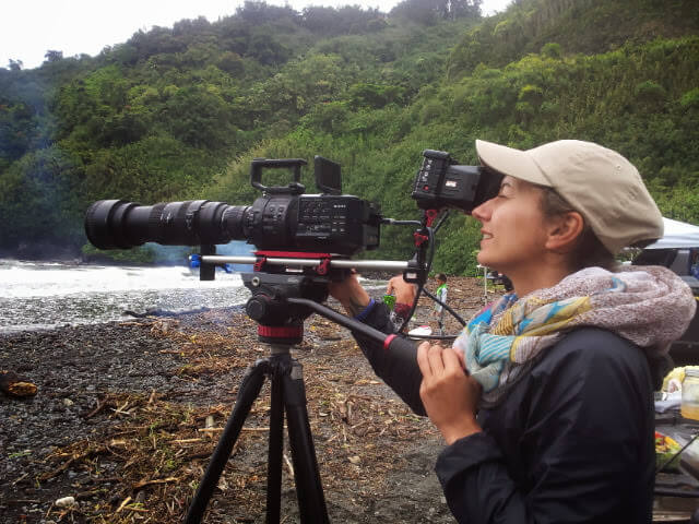 Production still from All of Our Heartbeats Are Connected Through Exploding Stars. A cameraperson looking to a lens of a camera on a beach.