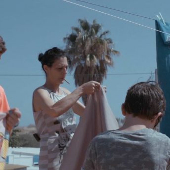 A still from The Boy and the Suit of Lights. A woman smoking a cigarette holds up a large piece of fabric. She stands in front of a palm tree and a little boy is in front of her, his back facing the camera.