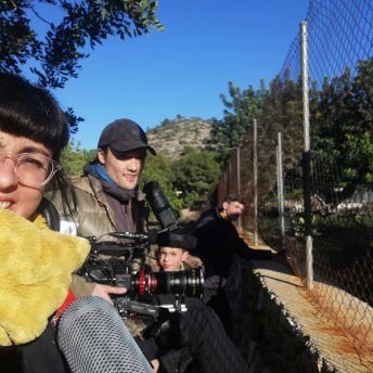 A production still from The Boy and the Suit of Lights. Inma de Reyes takes a selfie of herself and two crew members in front of a fence. One of the crew members holds a large camera.
