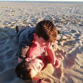 A still from The Boy and the Suit of Lights. Two young boys lie down on the sand. One is on top of the other. They are both smiling.