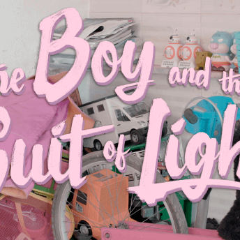 A photo of the graphic for The Boy and the Suit of Lights. The title is spelled out in pink cursive font over a pile of toys sitting in a white room.
