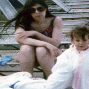 Photography of a woman sitting on a pool chair. In front of her is a toddler with a big white towel; the toddler is looking at the camera.